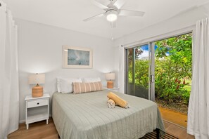 A bedroom with a bed, two nightstands, a ceiling fan, a window with a view of greenery, and a painting on the wall. - Kauai Puamana 4b 2 Bedroom Condo (Princeville)