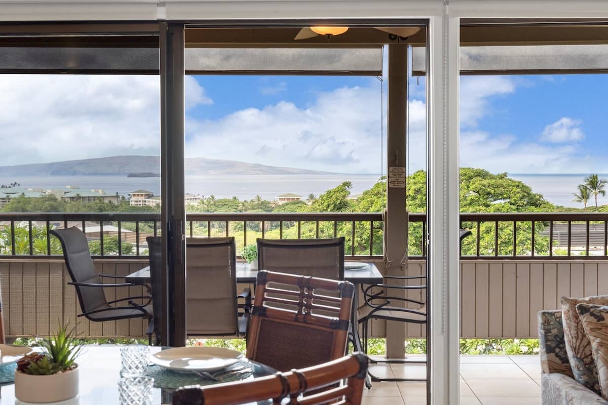 A covered patio with a table and chairs, overlooking a scenic view of a coastline and hills.