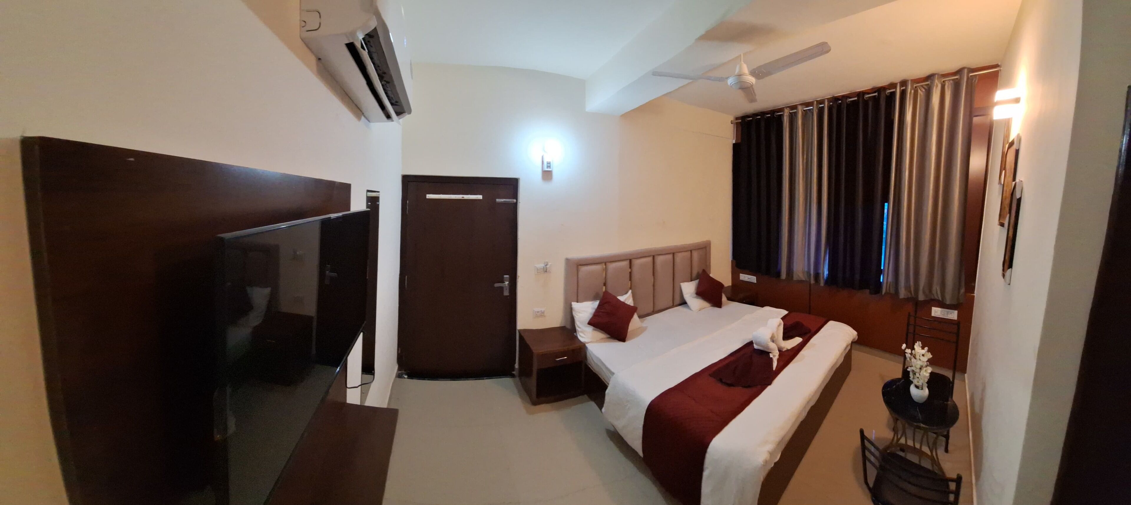 A hotel room with a bed, a TV, an air conditioner, a ceiling fan, and a window with curtains.