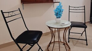 Une pièce dotée d’un téléviseur à écran plat fixé au mur, de deux chaises noires, d’une petite table ronde avec un plateau blanc et d’une plante artificielle bleue.