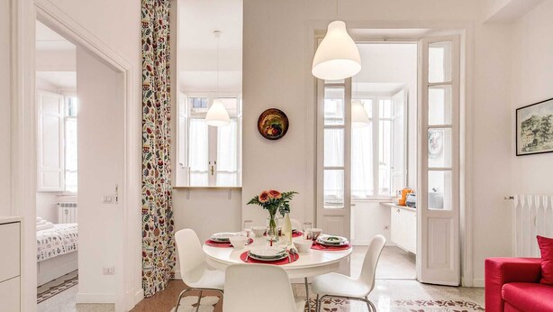 A dining area with a round table, white chairs, a vase with flowers, and a red couch.