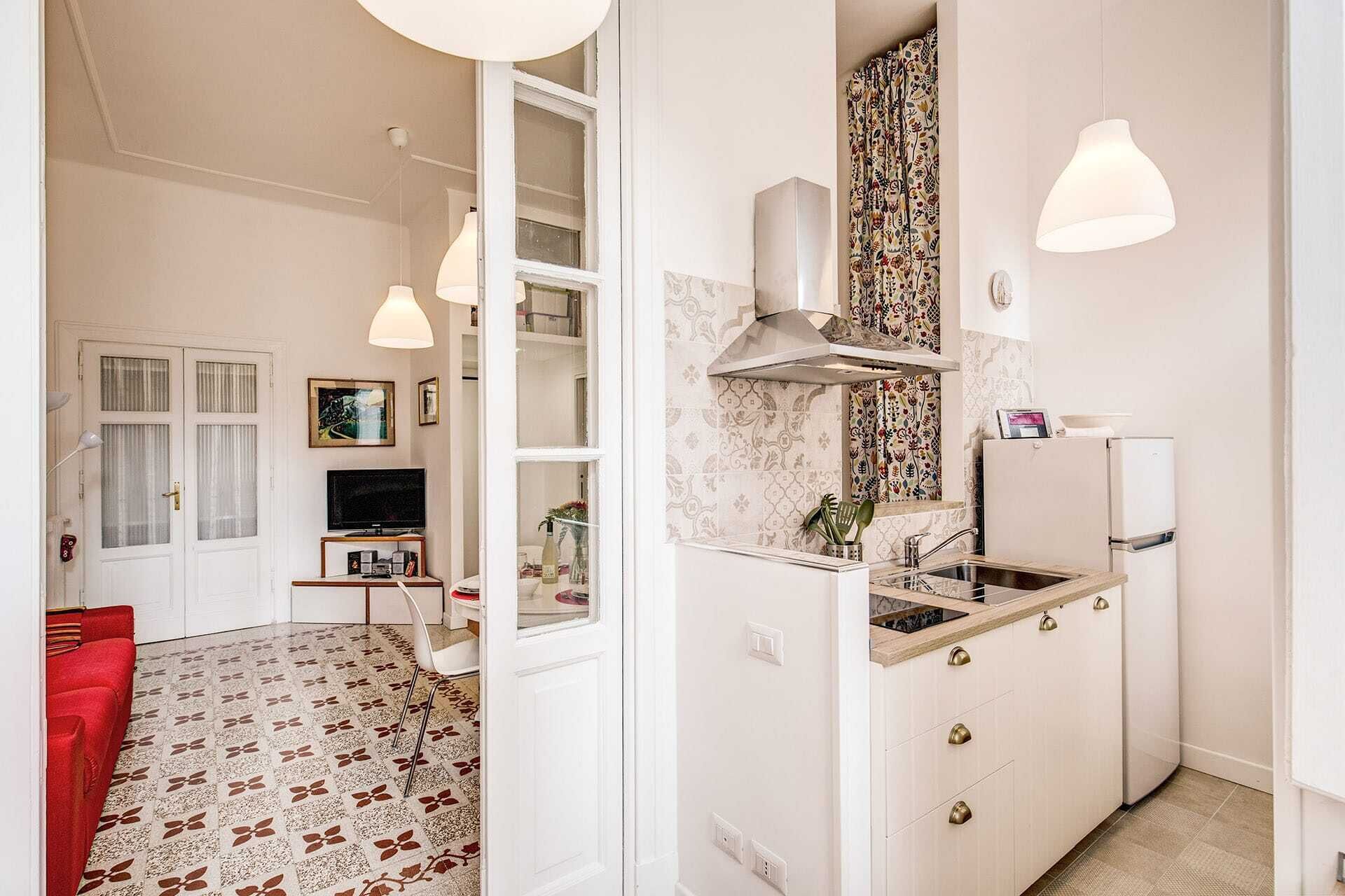 Cucina compatta con lavandino, fornello e piccolo frigorifero. Angolo ristoro con divano rosso e televisione. Pareti bianche e pavimento decorato.