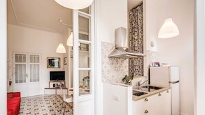 A compact kitchen with a sink, stove, and a small refrigerator. There is a dining area with a red sofa and a TV. The room has white walls and patterned flooring.