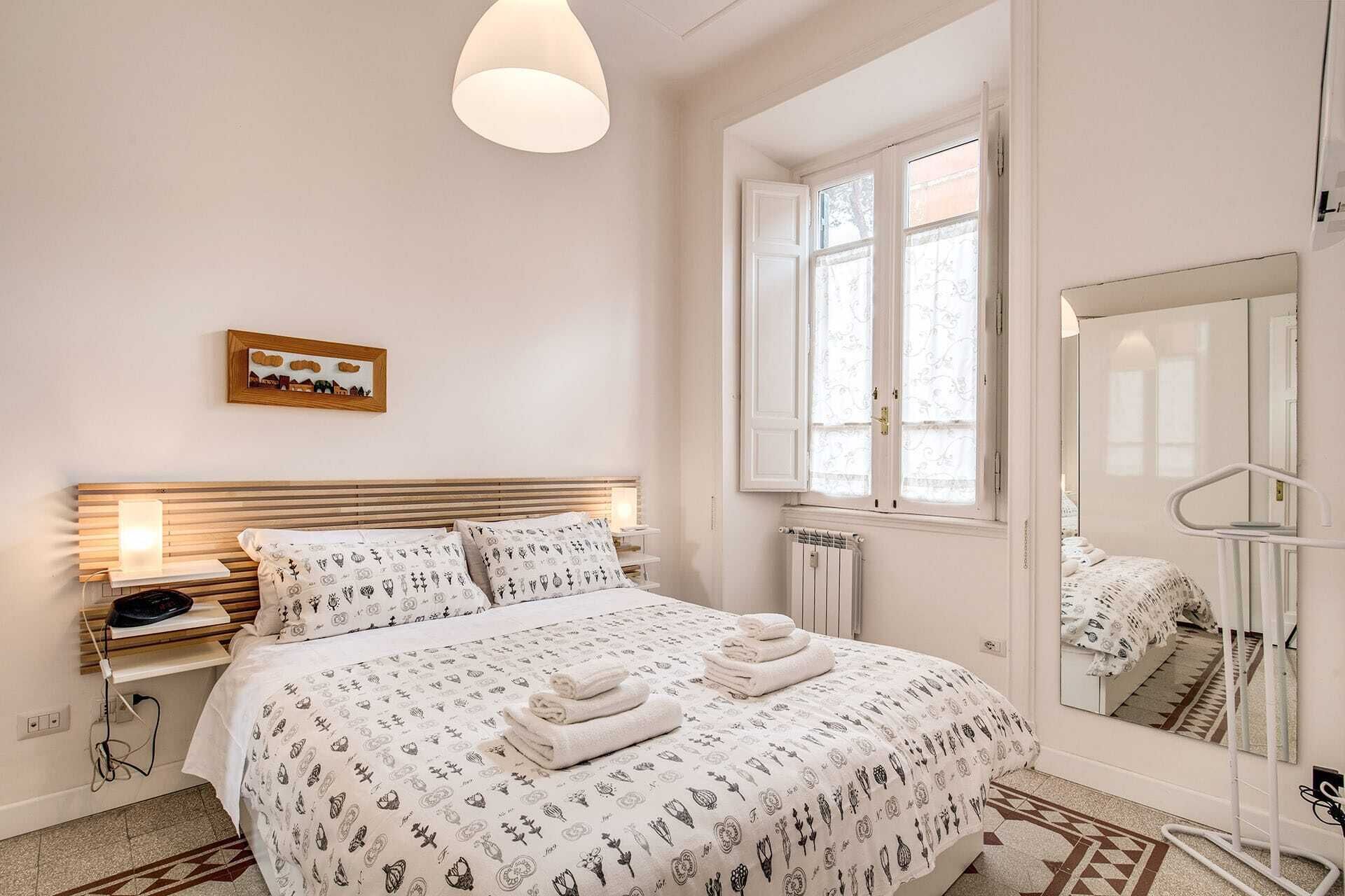 A bedroom with a bed, bedside table, lamp, window, and mirror.