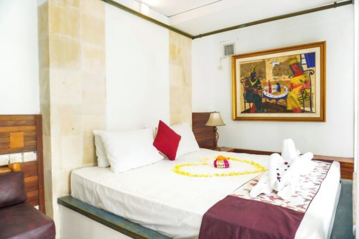 A hotel room with a bed, a red pillow, a white bedspread, a yellow flower arrangement, and a painting on the wall.
