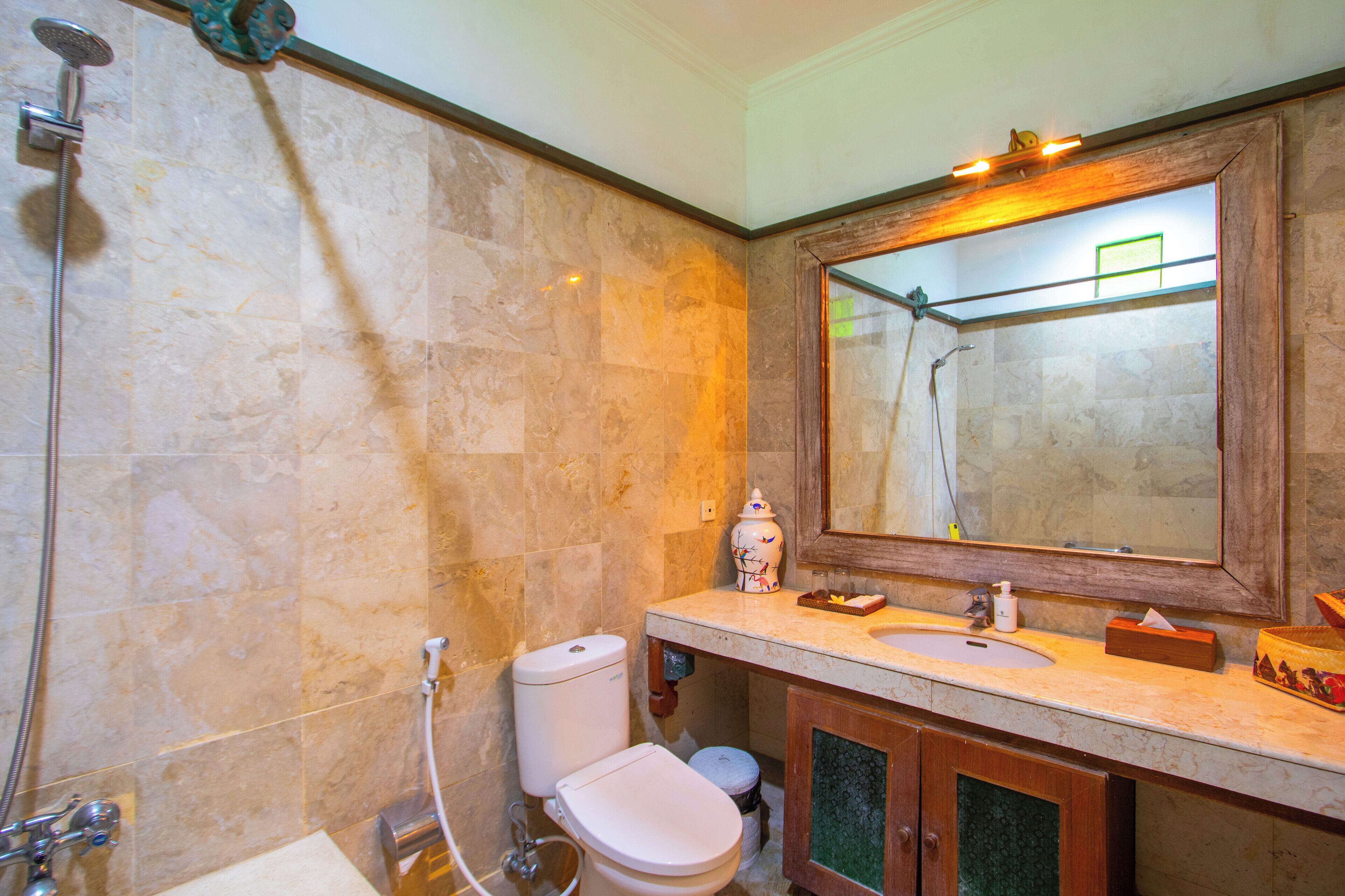 A bathroom with a marble wall, a wooden vanity, a large mirror, a toilet, and a shower.