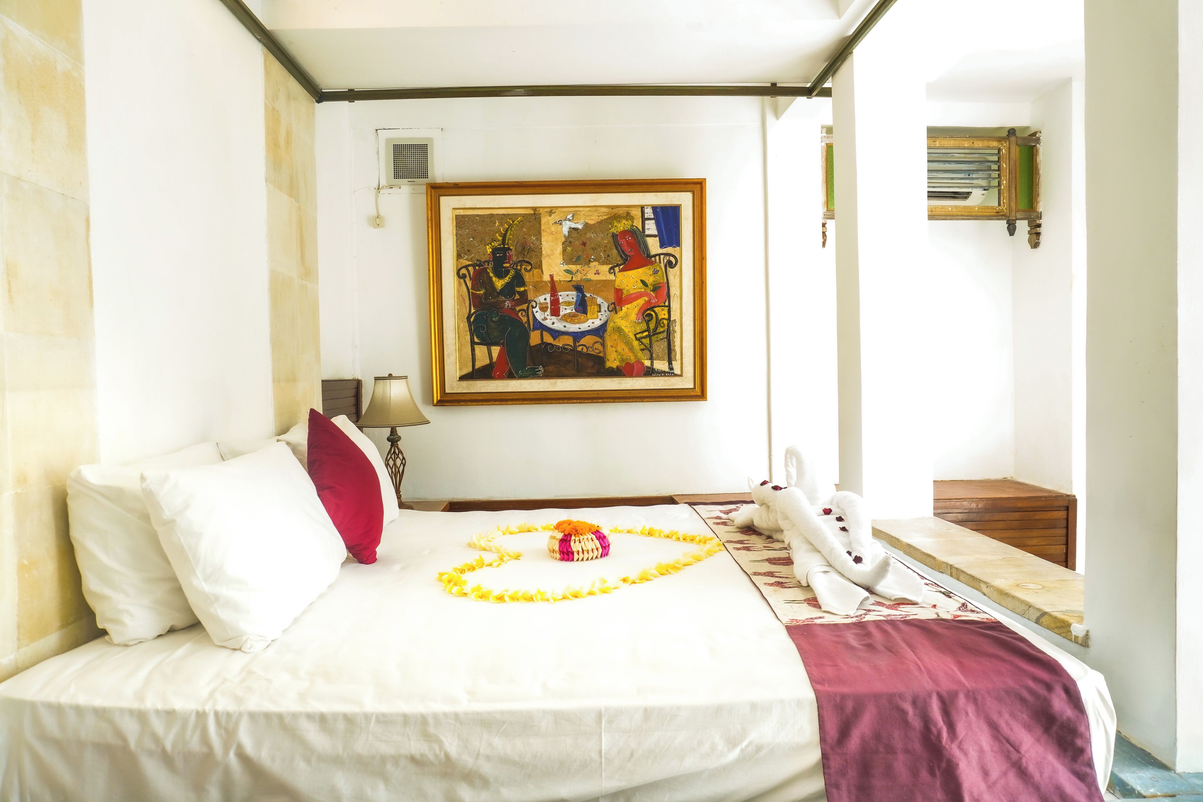 A hotel room with a bed, a painting on the wall, a small table with a flower arrangement, and a suitcase on the floor.