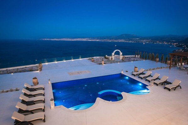 Una piscina infinita en la azotea con sillones de descanso, un arco decorativo y vistas a la costa al atardecer.