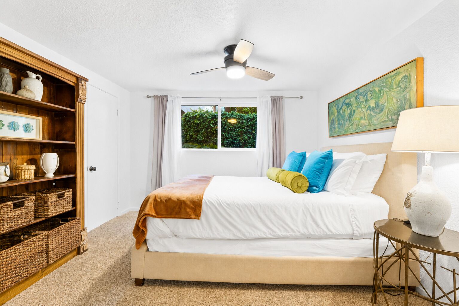 A bedroom with a bed, a ceiling fan, a window with curtains, a painting on the wall, and a side table with a lamp.