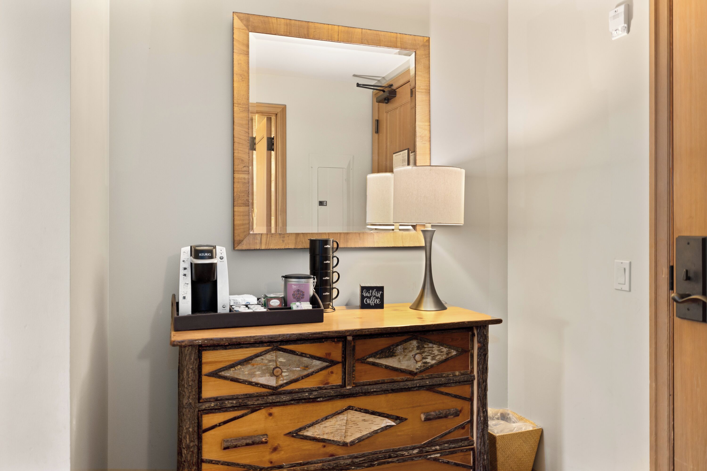 A wooden console table with a mirror above it, a lamp, a coffee maker, and a tray with mugs and a small sign.