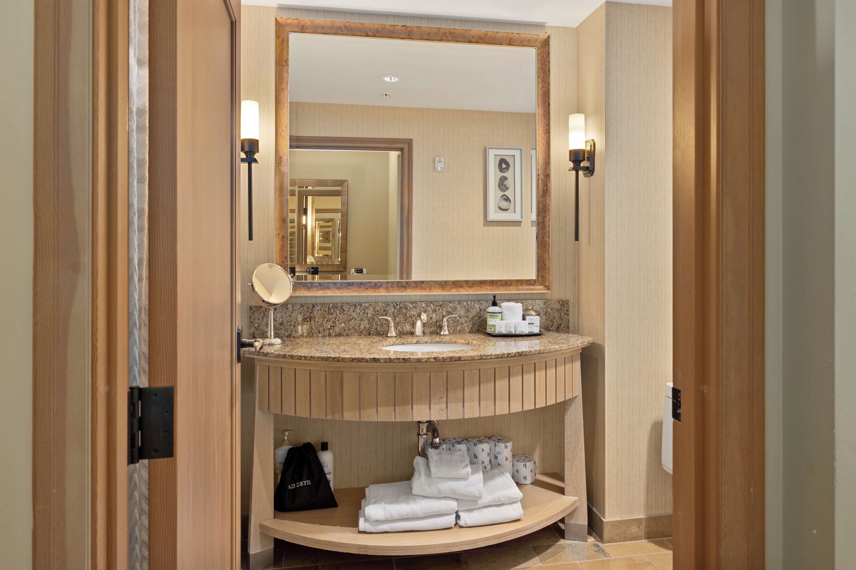 Une salle de bain d’hôtel avec un meuble-lavabo en bois, un grand miroir, deux appliques murales et un porte-serviettes.