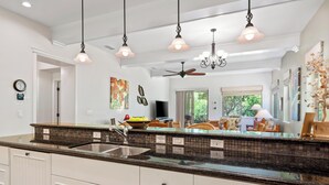 A modern kitchen with a large island, white cabinetry, and a black backsplash. There are pendant lights, a ceiling fan, and a window with a view of greenery.
