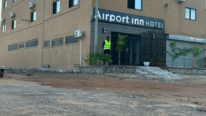 Ein mehrstöckiges Gebäude mit der Aufschrift „Airport Inn Hotel“. Es sind mehrere Klimaanlagen, ein Sicherheitsbeamter und eine gepflasterte Fläche vor dem Gebäude zu sehen.