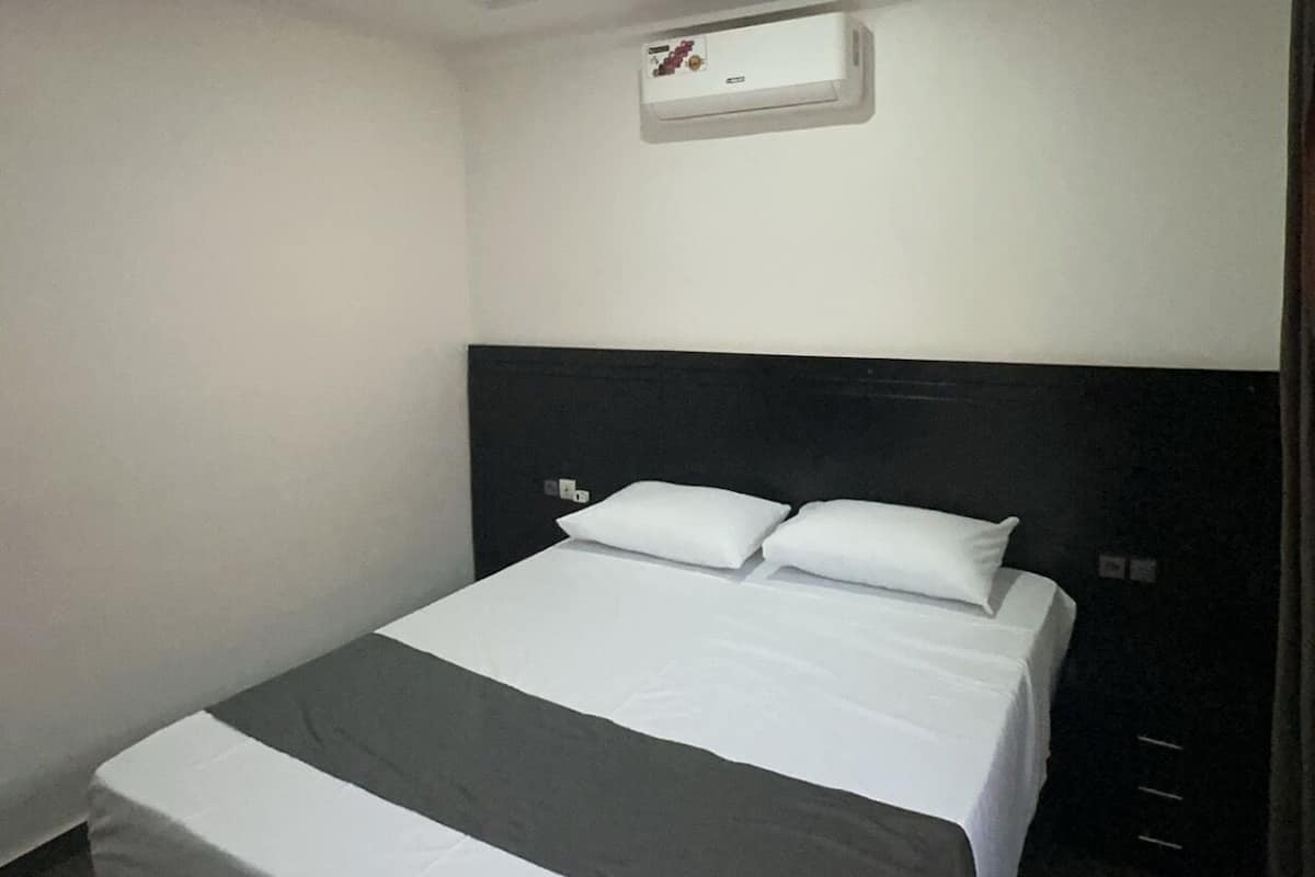 A hotel room with a bed, a nightstand, a wall-mounted air conditioning unit, and a door.
