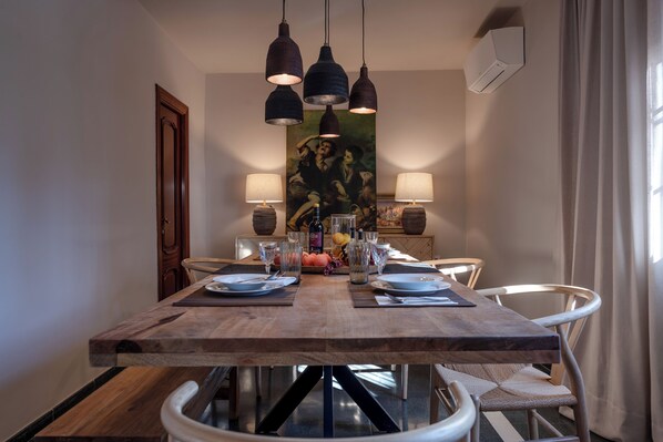 Een eetruimte met een houten tafel, vier stoelen, hangende吊り下げ灯, en een luchtverkoeler aan de muur.