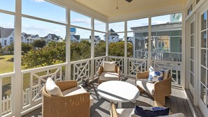 A covered outdoor area with wicker furniture, a ceiling fan, and large windows offering a view of houses and greenery.