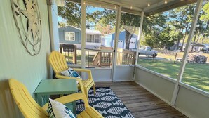A covered porch with yellow chairs and a table, a patterned rug, and a view of a residential area with houses and a truck. - Absolutely Darling private little oasis !  (Holly Ridge)