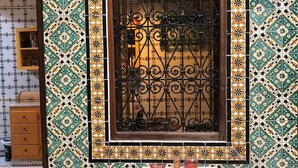 A room with a patterned tile wall, a window with decorative iron bars, a small table with a cushion, and two stools. - Atelier D (Tunis)