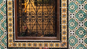 A room with a window featuring ornate black iron bars, surrounded by a tiled wall with a geometric pattern. There is a small table with a white top and a red cushion, accompanied by two green stools. - Atelier D (Tunis)