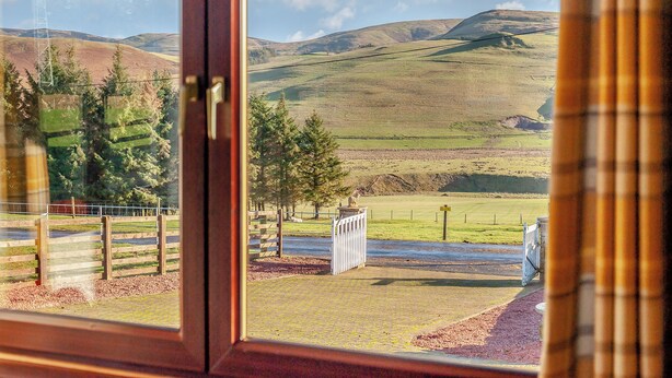 A view through a window of a rural landscape with rolling hills, a body of water, and a fenced area with trees. - Towford Farm Bungalow (Jedburgh)