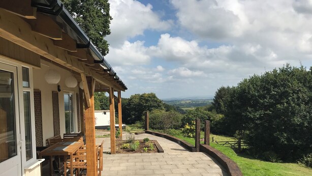 A covered patio with a view of the countryside, wooden furniture, and a paved walkway. - Quiet and secluded farmhouse, with far reaching views, spacious and comfortable  (Liskeard)
