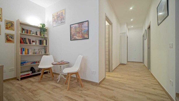 Ein Flur mit Holzboden, weißen Wänden und einem Bücherregal mit Büchern und einer Pflanze. Zwei weiße Stühle und ein kleiner runder Tisch stehen dort.