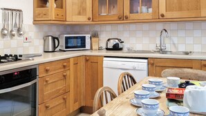 A kitchen with wooden cabinets, a microwave, a kettle, a dishwasher, and a dining table set with blue and white cups.