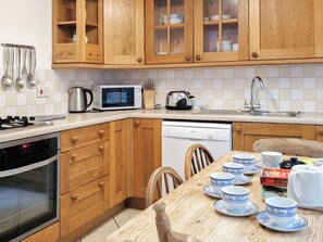 A kitchen with wooden cabinets, a microwave, a kettle, a dishwasher, and a dining table set with blue and white cups.