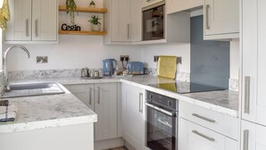 A modern kitchen with white cabinetry, a built-in oven, and a countertop with various appliances. - 2 bedroom accommodation in Criccieth (Criccieth)