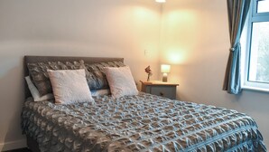 Un lit bien fait, avec une couvre-lit à motifs et des oreillers, une table de chevet avec une lampe, et une fenêtre avec des rideaux.