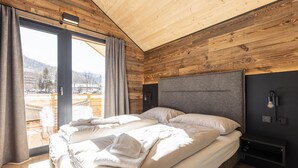 Une chambre avec un plafond en bois, un grand lit recouvert de draps blancs, une table de chevet et une vue sur un paysage enneigé par une porte coulissante.