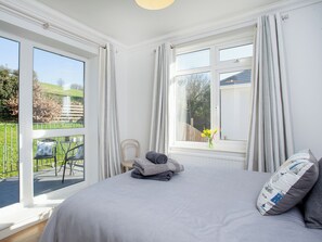 Un dormitorio con un ventanal grande, una cama con manta gris, una silla y una mesita.
