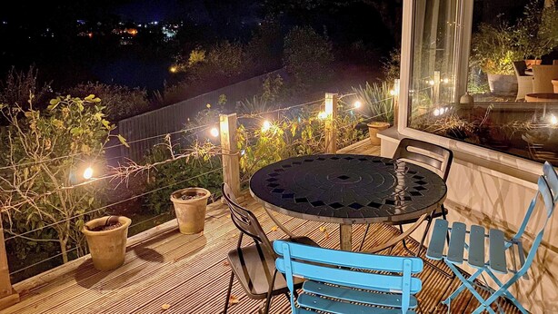 Een terras buiten met een ronde tafel en blauwe stoelen, omringd door potplanten en lichtslingers.