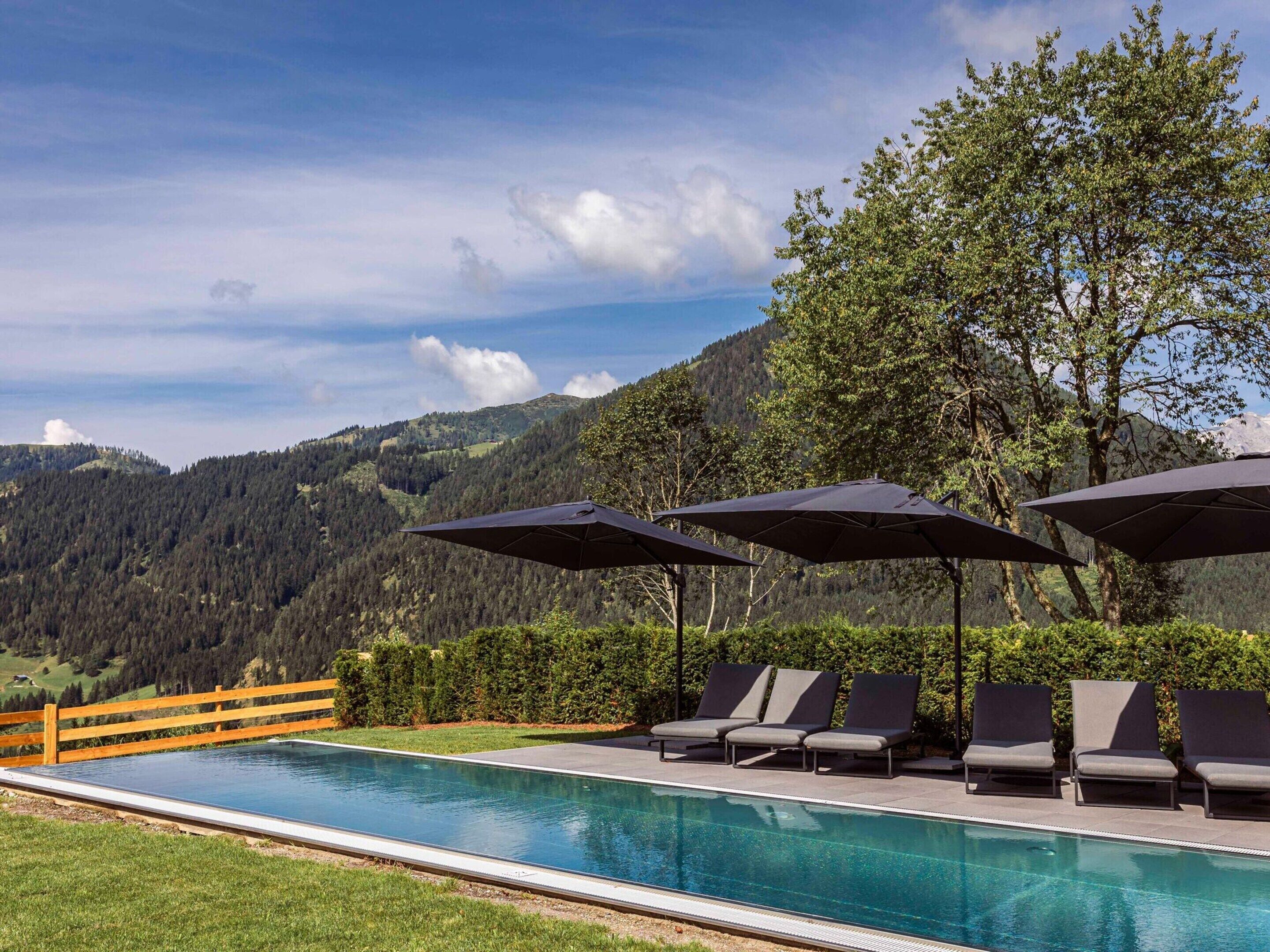 A swimming pool with lounge chairs and umbrellas, surrounded by a wooden fence and trees, with a mountainous backdrop.