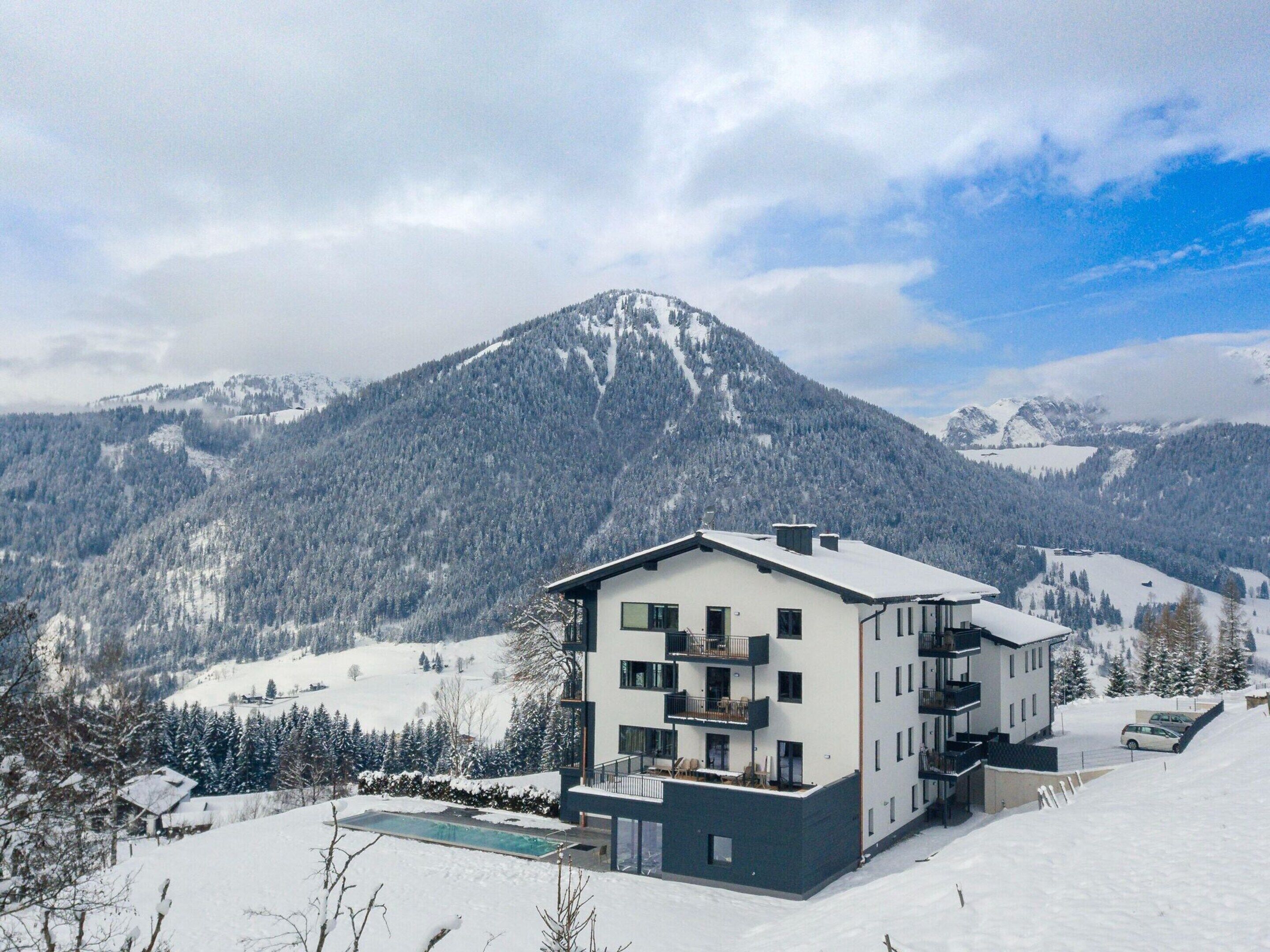 A modern multi-story building with balconies, surrounded by snow-covered mountains and trees.