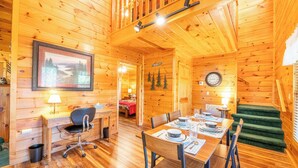 A wooden cabin interior with a dining area, a desk with a chair, a staircase, and a bed in the background.