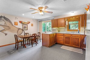 A kitchen with wooden cabinets, a dining table with chairs, a ceiling fan, and a window with a view of trees. - Charming 3-bedroom house with AC and WiFi on Sandy Pond. (Sandy Creek)