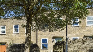 A stone building with multiple windows, a tree in the foreground, and a stone wall. - The Cottage on the Green - UK47990 (Chipping Norton)