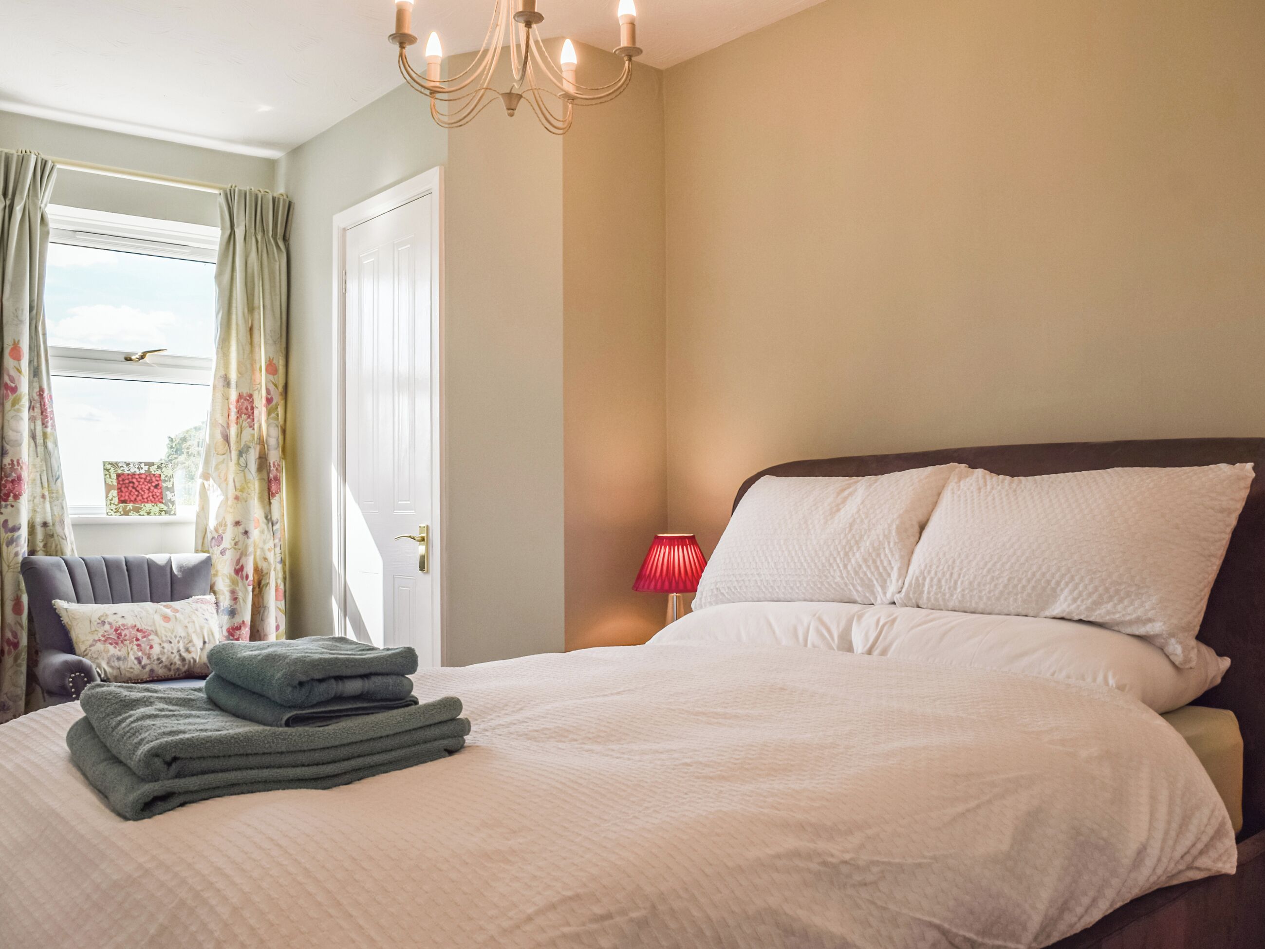A bedroom with a bed, a chair, a window with curtains, and a chandelier.