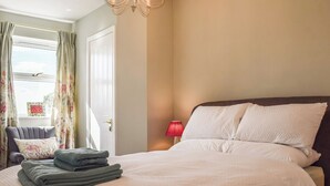 A bedroom with a bed, a chair, a window with curtains, and a chandelier. - The Cottage on the Green - UK47990 (Chipping Norton)