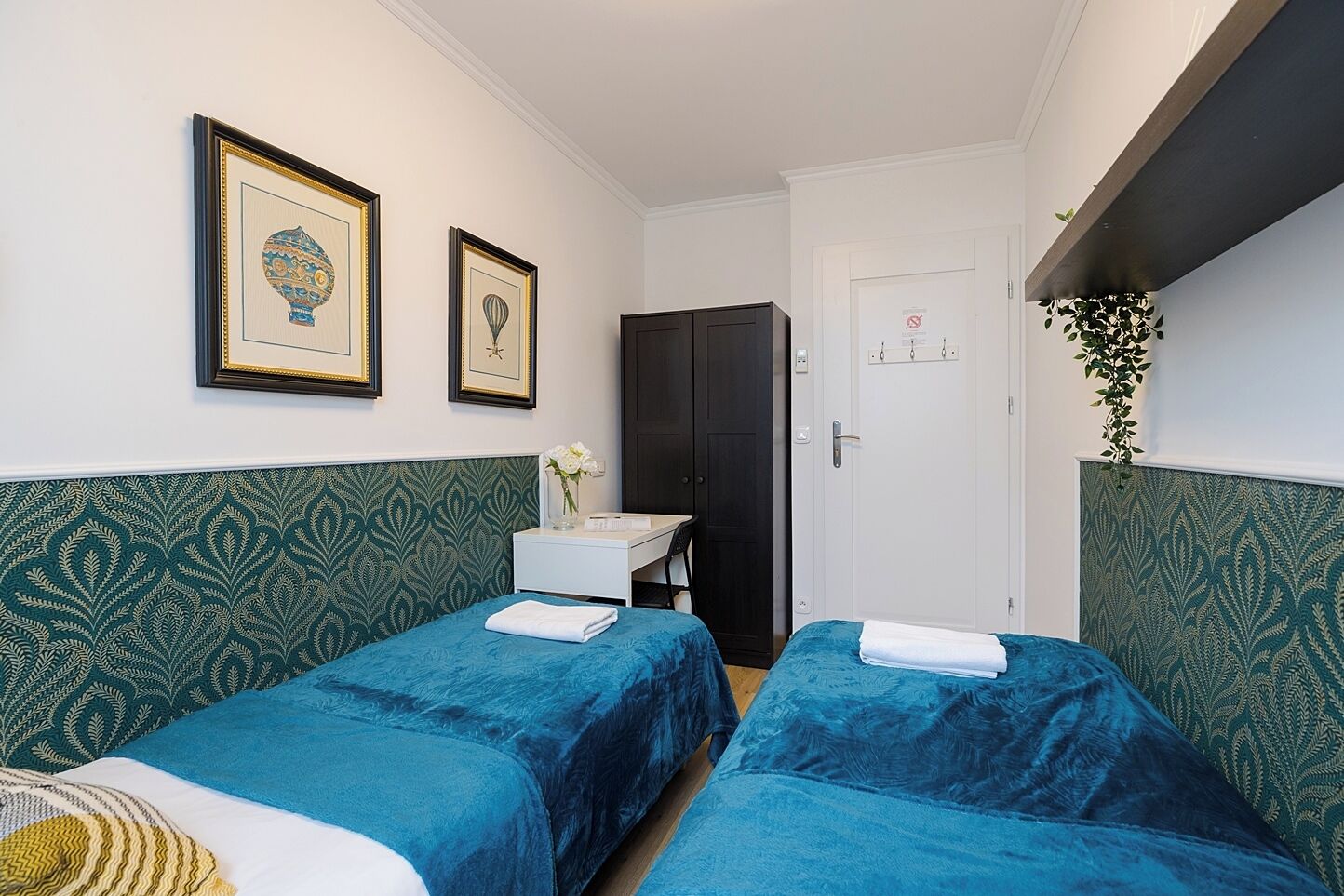 Une chambre d’hôtel avec deux lits, un petit bureau, un lavabo blanc et des œuvres d’art encadrées accrochées au mur.