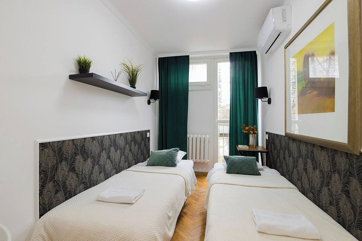 A hotel room with two single beds, a small table with a vase of flowers, a shelf with plants, and a window with curtains.