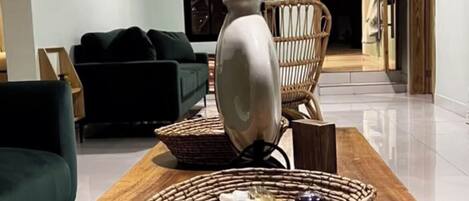 A living room with a wooden coffee table, a white vase, and a wicker basket with stones. There is a green sofa and a wicker chair in the background.
