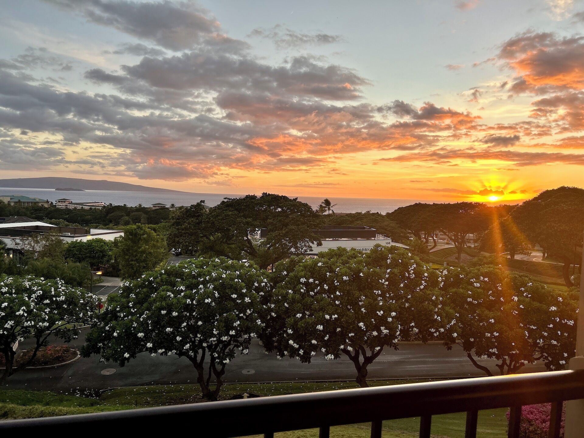 Vista do pôr do sol a partir de uma varanda com vista para um estacionamento, árvores e um corpo de água ao longe.