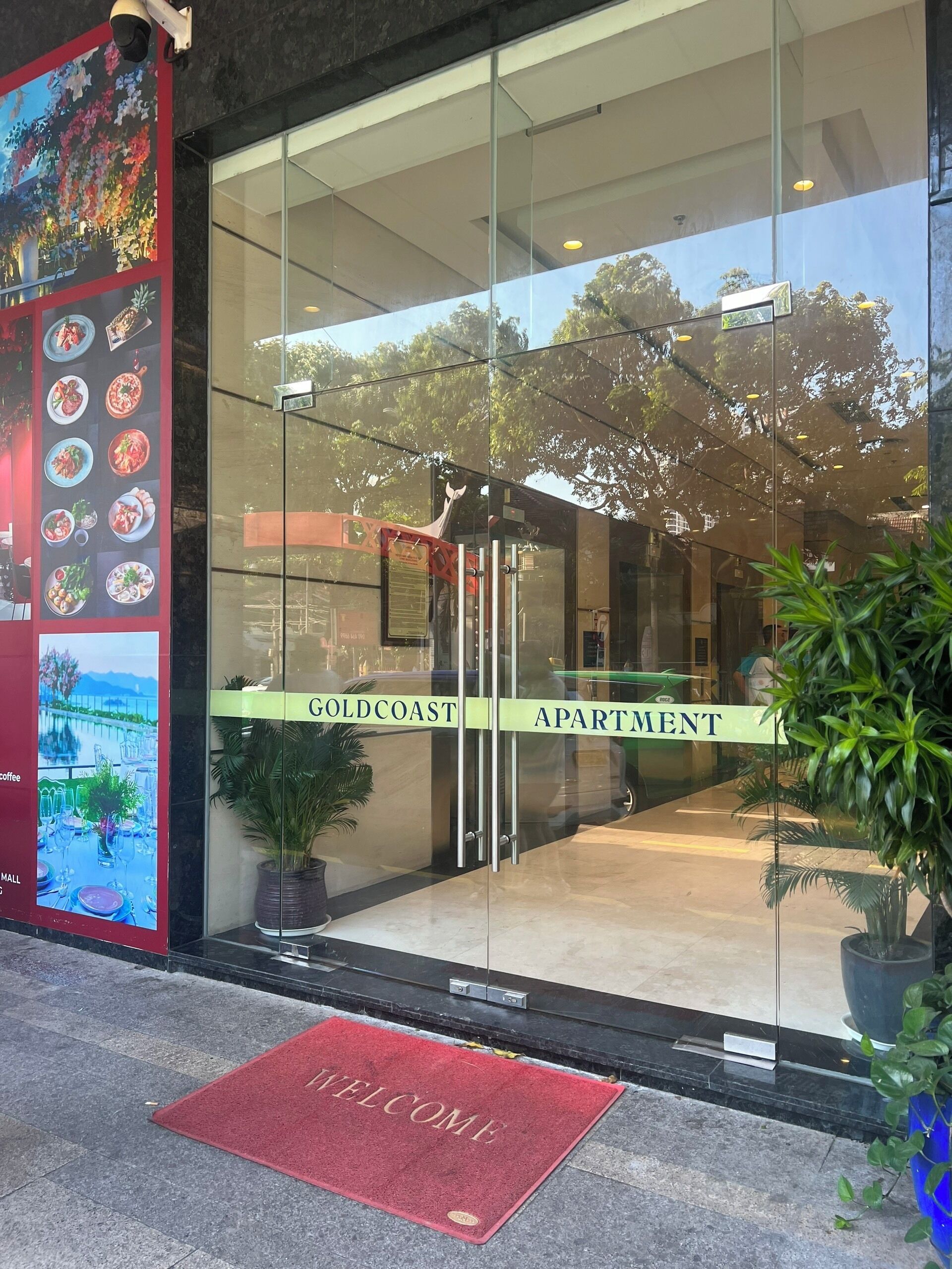 Ingresso di un edificio moderno con facciata in vetro, tappeto di benvenuto rosso e insegna che recita 'Goldcoast Apartment'.