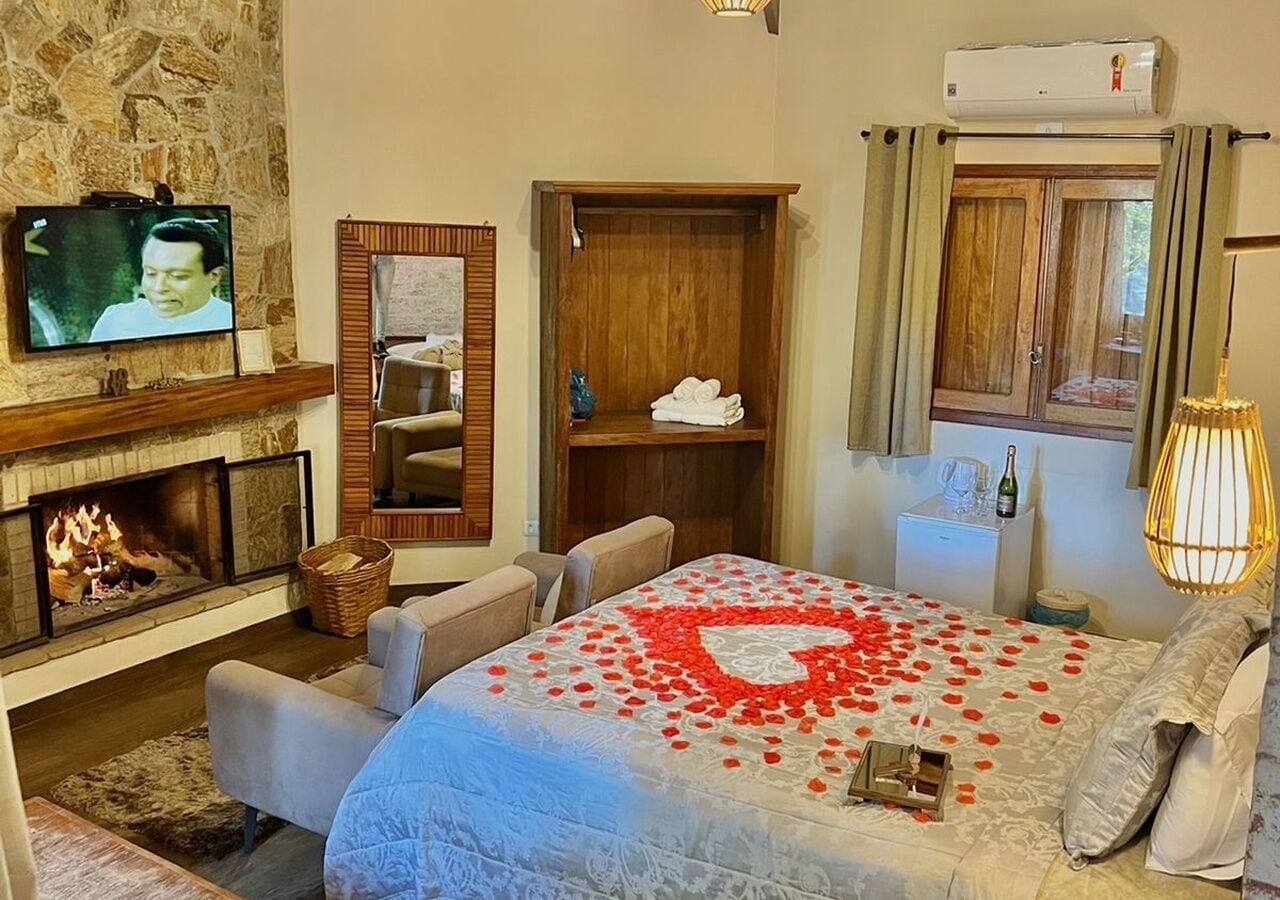 A hotel room with a fireplace, a television, a bed with rose petals, a wooden wardrobe, and a window with curtains.