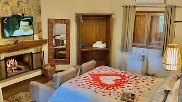 Habitación de hotel con chimenea, televisor, cama con pétalos de rosa, armario de madera y ventana con cortinas.