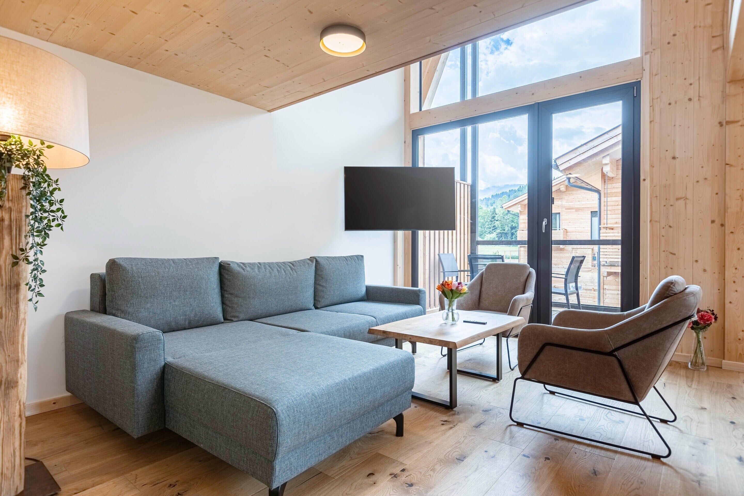 Una sala de estar moderna con un sofá seccional, una mesa de centro y un sillón reclinable. Un televisor de pantalla plana está montado en la pared, y hay un ventanal que ofrece vistas al exterior.