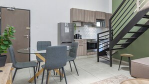 A modern kitchen with a stainless steel refrigerator, wooden cabinets, and a built-in oven. There is a dining area with a glass table and grey chairs. A staircase with a black railing leads to an upper level.