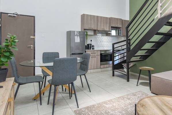 A modern kitchen with a stainless steel refrigerator, wooden cabinets, and a built-in oven. There is a dining area with a glass table and grey chairs. A staircase with a black railing leads to an upper level.